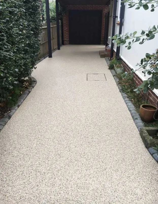 Cream resin bound side driveway with charcoal sett edging running beneath a carport to a garage
